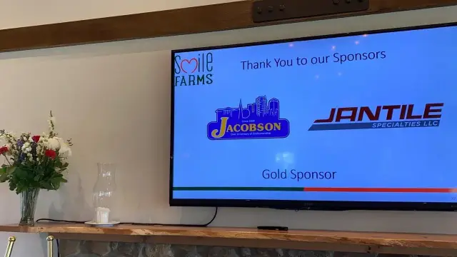 TV screen displaying Smile Farms sponsors, including Jacobson and Jantile Specialties LLC, labeled as Gold Sponsor, with flowers on a shelf nearby.