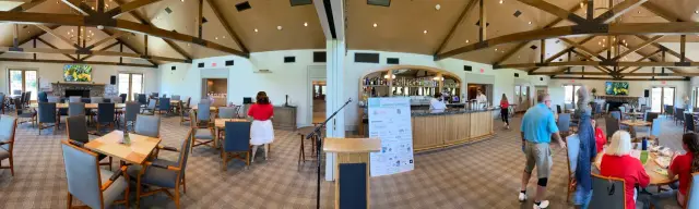 Panoramic view of a spacious, bright dining area with wooden tables and chairs, high vaulted ceiling with exposed beams, a bar on one side with people standing nearby, and a fireplace with a TV above it in the background.