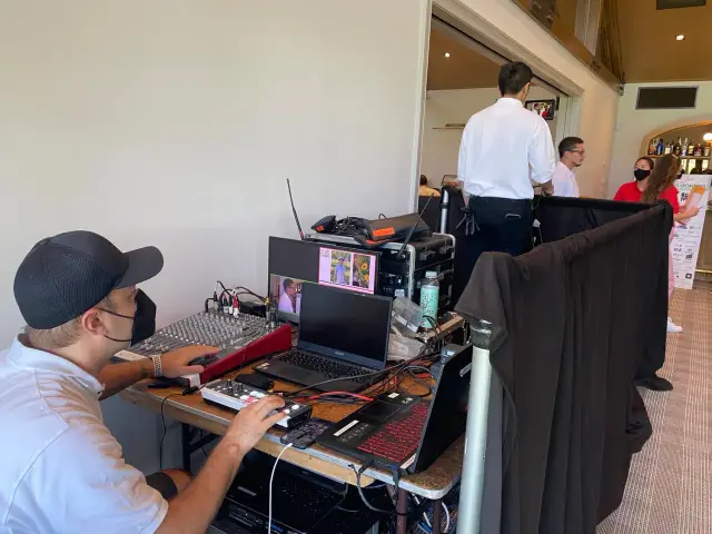 A person operating audio and video equipment with multiple screens and a mixing console, wearing a black cap and mask. Several people are standing nearby, some wearing masks, in a room with a setup for event production.