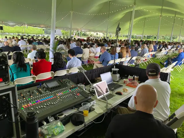 Outdoor event under a large tent with attendees seated on folding chairs, seen from an audio-visual control station with mixing console and laptops in the foreground.