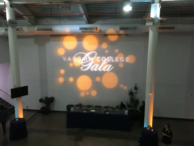 Indoor event with 'Vaughn College Gala' projected on a wall, partial view of a reception area with a bar, decorative lighting, and a few people present.