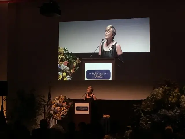A person speaking at a podium during the Vaughn College Gala, with a large screen displaying the speaker's image behind them. The stage is decorated with flowers.