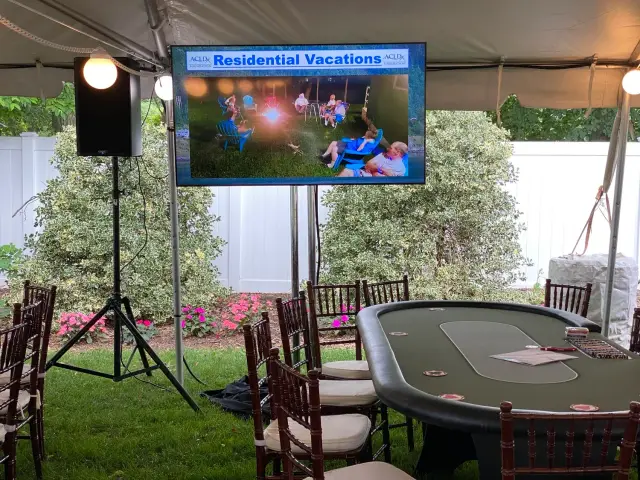 Outdoor event setup with a poker table, chairs, and a screen displaying a "Residential Vacations" slide under a tent.