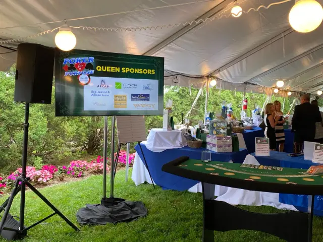 Outdoor event under a tent with a "Poker for Purpose" sign, sponsor logos, and a blackjack table. People are socializing near gift baskets on tables.