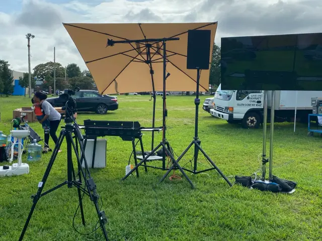 Outdoor setup with camera equipment, tripod, large umbrella, monitor, speaker, and people at an event in a grassy area.