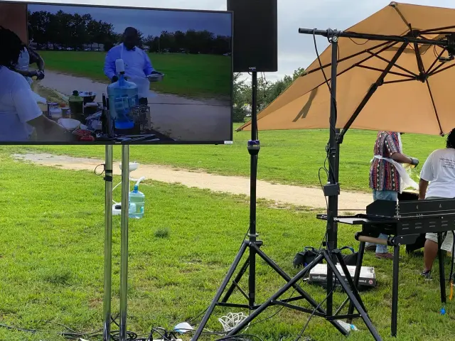 Outdoor event featuring a cooking demonstration setup. A large monitor displays a video of a person cooking outdoors on a table with various kitchen tools and ingredients.