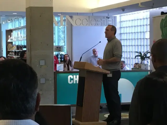 A man speaking at a podium in a bookstore-like setting, with shelves of products visible in the background and a small audience watching.