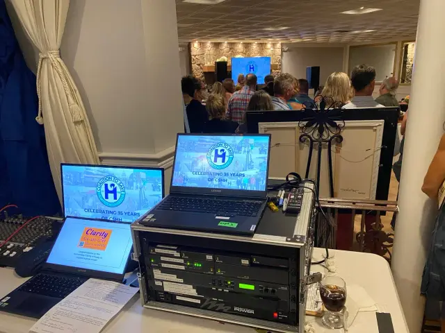 Event sound control area with laptops displaying "Celebrating 35 Years of CSHH" logo, surrounded by audio equipment, mixer, and a drink, with a crowd in the background at a gathering.