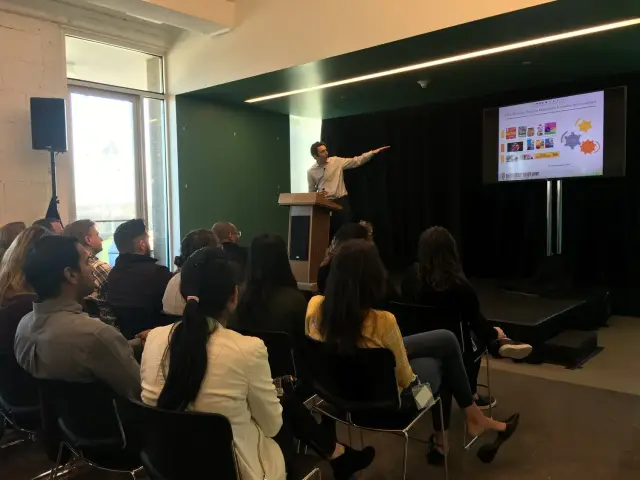 A person standing behind a podium giving a presentation to an audience seated in a room. The speaker is pointing to a slideshow displayed on a screen. The audience is attentively listening in a modern conference space with large windows.