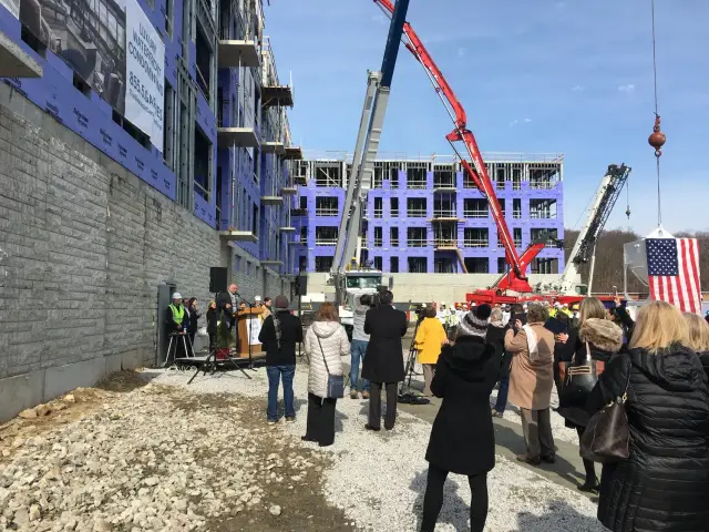 A construction site with a building partially completed, surrounded by people attending an event. The building has blue sheathing, and cranes are visible. An American flag is displayed on a structure being lifted by a crane.