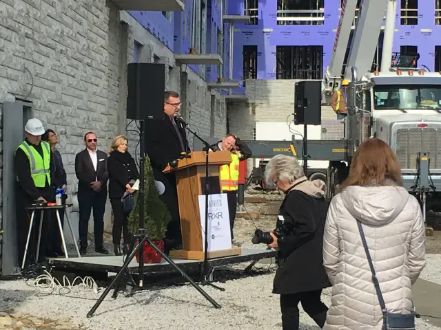 A man speaks at a podium during a construction site event. Surrounding the podium are people in coats, including a photographer with a camera, a worker in a safety vest and hard hat, and a large truck.