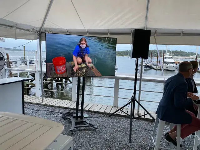 Outdoor event setup with a large screen displaying a girl working with a bucket near water. The event takes place under a canopy near a marina with boats. Attendees sit at tables.
