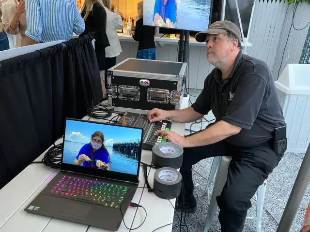 Man with a cap operating audio-visual equipment at an outdoor event, with a laptop displaying a person wearing a mask by a waterfront on the screen. Multiple cables and a mixing console are visible. People in the background are standing near a bar.