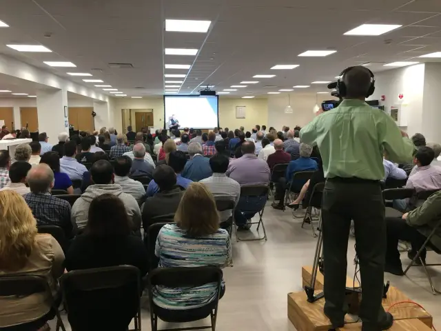 A large audience seated in a conference room with a projector screen at the front. A person is filming the event with a video camera on a tripod.