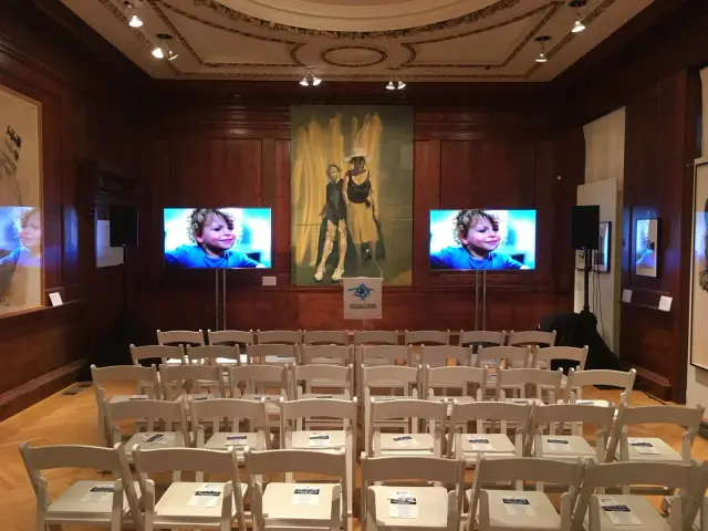 Room with rows of white chairs facing two large screens displaying an image of a child, paintings on the walls, and a lectern at the front.