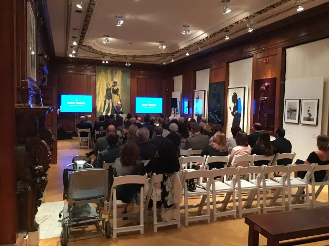 Audience seated in an art gallery with framed artworks on the walls, listening to a speaker at a podium.