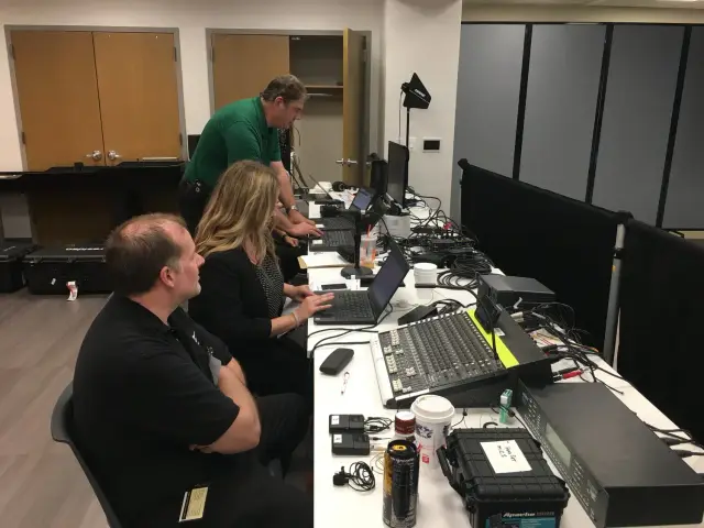 People working at a tech setup with laptops, sound equipment, and cables on a table in an office environment.