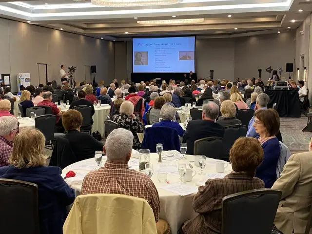 A conference room filled with people seated at round tables, watching a presentation on a large screen. The audience is attentively facing the presenter at the front, where visuals are projected.