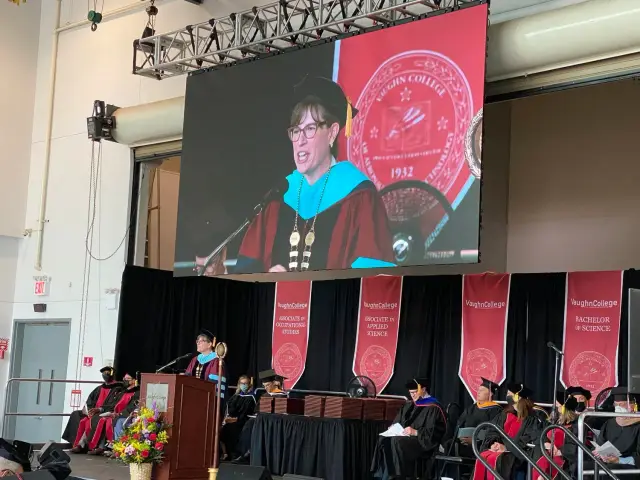 Commencement ceremony at Vaughn College with a person speaking at a podium on stage with banners and faculty seated. A large screen displays the speaker.