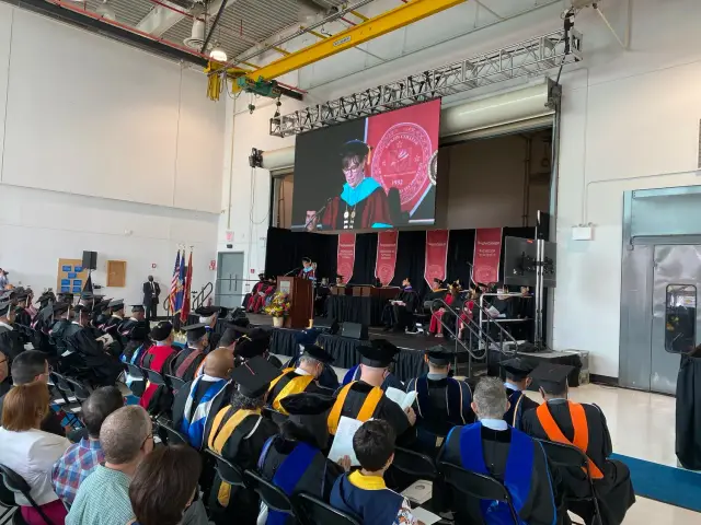 A college graduation ceremony taking place in an indoor auditorium. Graduates wearing caps and gowns sit in rows facing the stage. A podium is set up on the stage with a person delivering a speech.