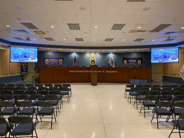 Empty meeting hall with chairs facing a stage at the International Brotherhood of Teamsters Local 282, featuring banners and video screens.