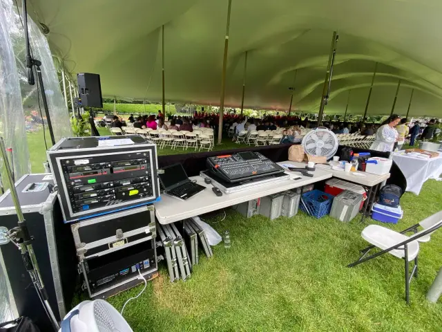 Outdoor event under a large tent with audio equipment and seating area, people gathered.