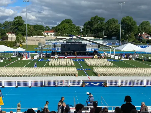 Outdoor graduation ceremony setup with stage, chairs, and tents on a sports field.
