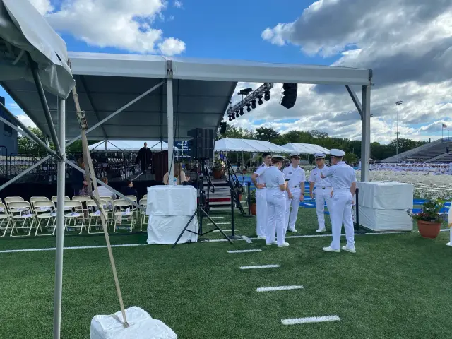 Military personnel in white uniforms standing near a stage and seating area on a grassy field, under a large tent, with a seated audience and speaker in view.