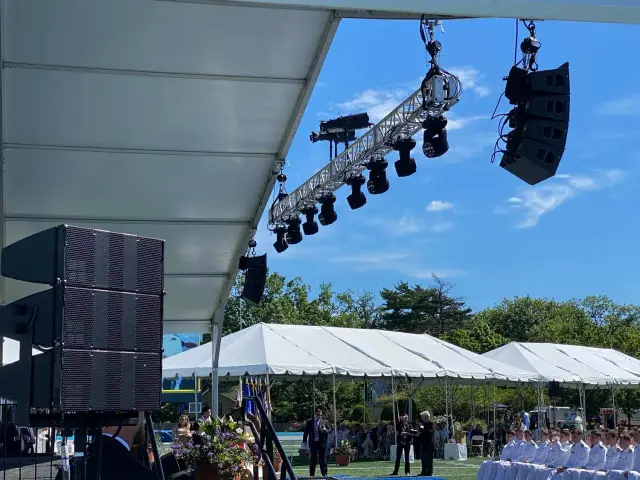 Outdoor event setup with large speakers and stage lighting under a tent, rows of seated people in white uniforms, and white tents in the background.