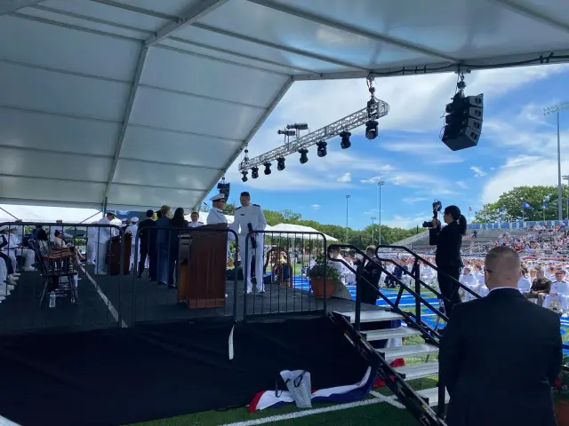 Graduation ceremony with people in formal military attire on a stage under a large tent, a photographer capturing the event, and an audience seated in bleachers at an athletic field.