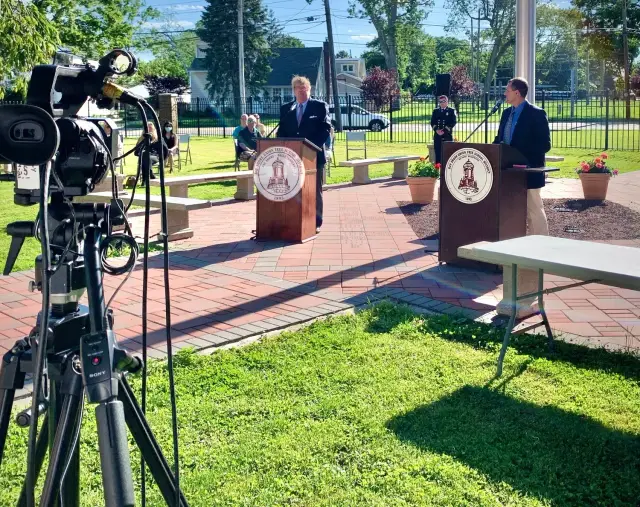 Two people speaking at podiums outdoors with a camera recording them, surrounded by a small audience seated on benches. The event is held on brick paving with a green lawn and trees in the background under clear blue skies.