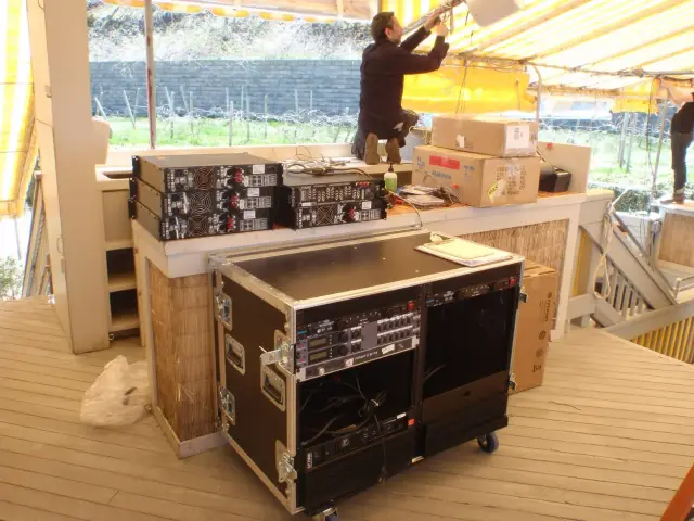 Outdoor setup with audio equipment, amplifiers, and people adjusting lighting under a yellow canopy.