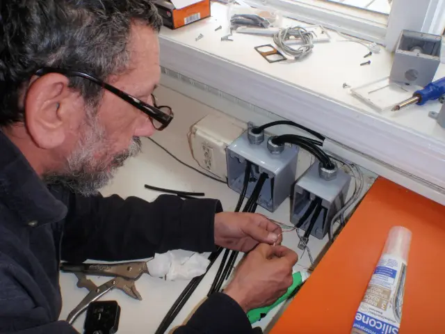 A person working on electrical wiring, connecting wires in electrical boxes, with tools and supplies on the table.