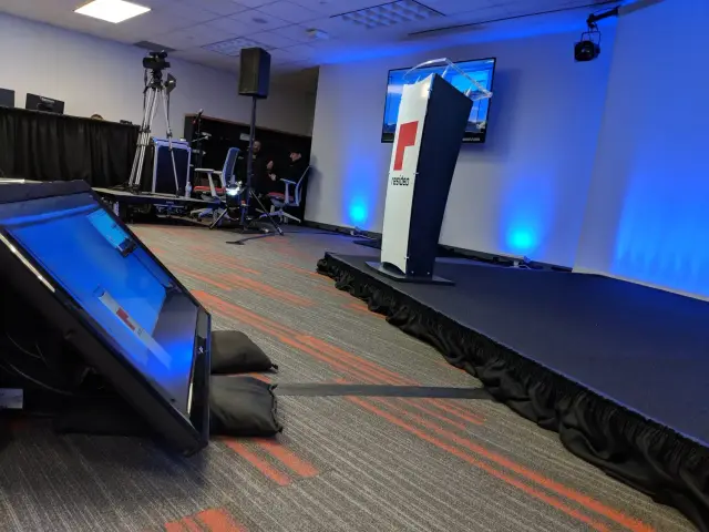 Empty stage setup with podium, video equipment, and a large monitor in a conference room.
