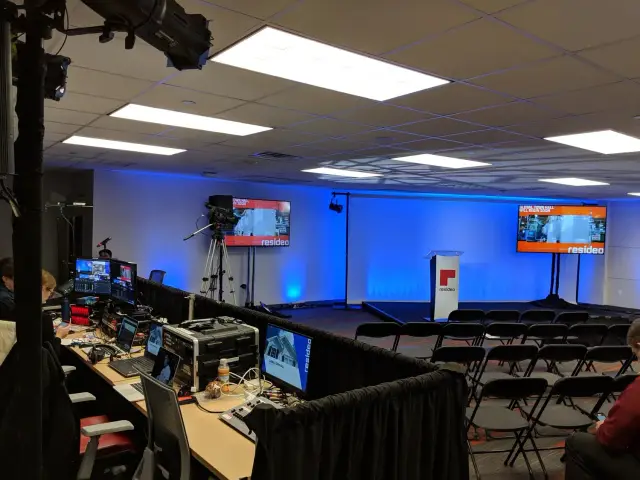 Conference room setting with a stage, podium, and chairs. There are multiple screens displaying the "Resideo" logo. Camera equipment and a control table with monitors are set up for recording or live streaming.