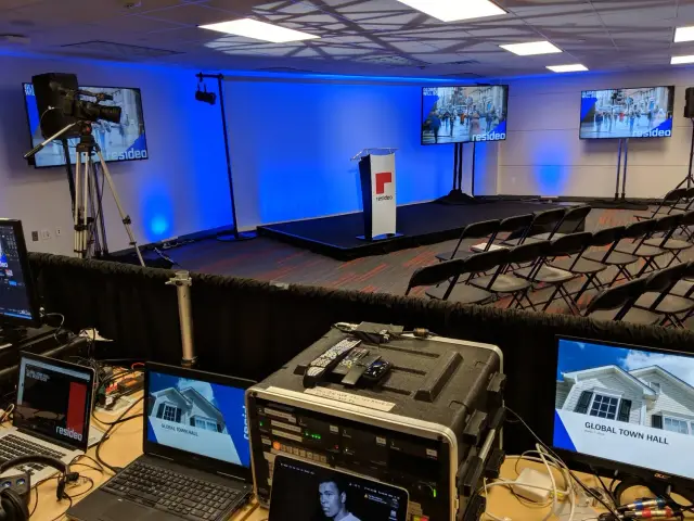 Empty conference room with stage and audio-visual equipment setup. Rows of empty chairs face a podium and multiple large screens. Technical equipment, laptops, and cables are visible in the foreground.