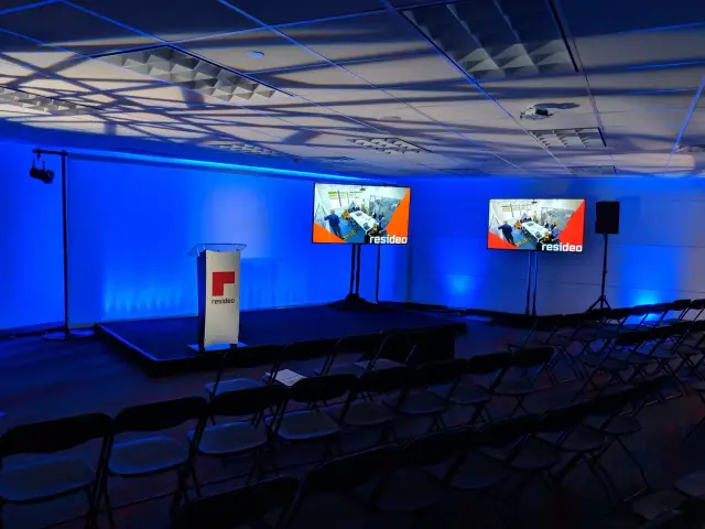 A conference room set up with rows of chairs facing a stage, featuring a podium with the 'resideo' logo, and two large screens displaying the same image. The room is illuminated with blue lighting.