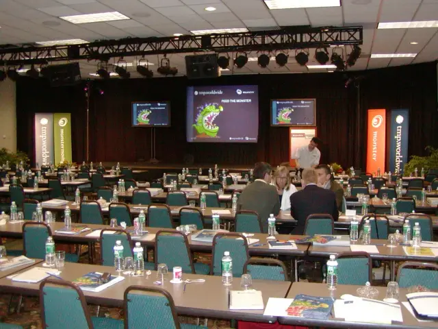 Conference room setup with chairs and tables, screens on stage, and branded banners, prepared for a presentation or meeting. There are water bottles and notepads on the tables, and a few people are discussing at the front.